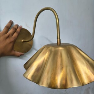 May include: A wall-mounted lamp featuring a brass-toned finish with a curved arm and a wide, scalloped, petal-shaped shade. The fixture includes a circular wall plate and a downward-facing, fluted metal lampshade with a metallic sheen.