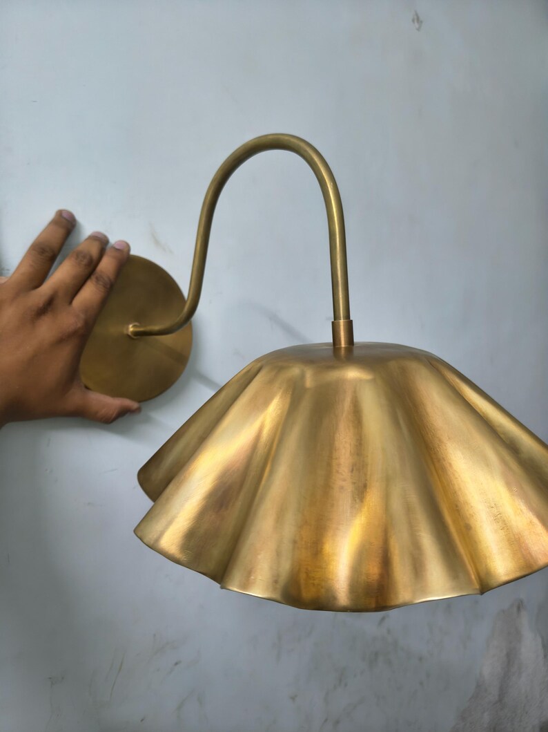 May include: A wall-mounted lamp featuring a brass-toned finish with a curved arm and a wide, scalloped, petal-shaped shade. The fixture includes a circular wall plate and a downward-facing, fluted metal lampshade with a metallic sheen.