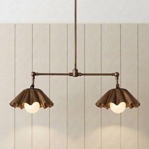 May include: A bronze-toned metal pendant light fixture featuring a central horizontal rod suspended from a single vertical stem. Two downward-facing, ruffled, bell-shaped shades are attached to the ends, each housing a round, white glass bulb.