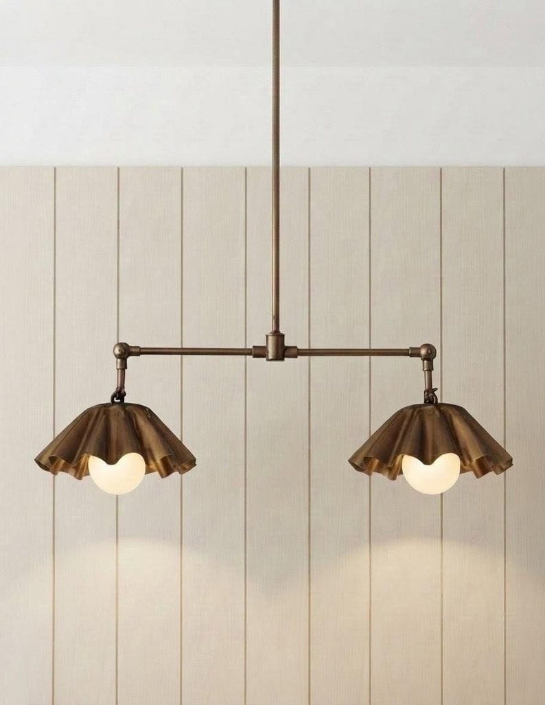 May include: A bronze-toned metal pendant light fixture featuring a central horizontal rod suspended from a single vertical stem. Two downward-facing, ruffled, bell-shaped shades are attached to the ends, each housing a round, white glass bulb.