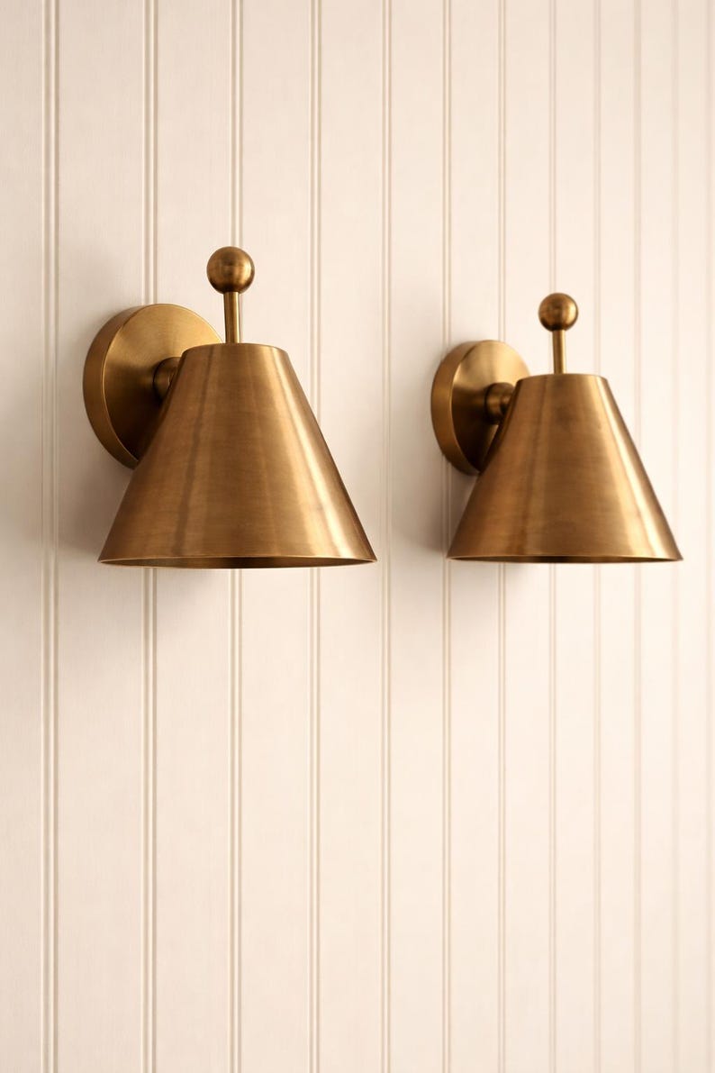 May include: Two brass sconces with a conical shade and a round backplate are mounted on a white, vertical-paneled wall. Each sconce has a small, spherical detail at the top. The sconces have a warm, metallic finish.