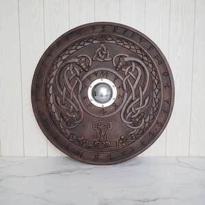 May include: A round, brown wooden shield with intricate carvings. The shield features a central silver boss, knotwork designs, and runes around the edge. A hammer symbol is carved at the bottom.