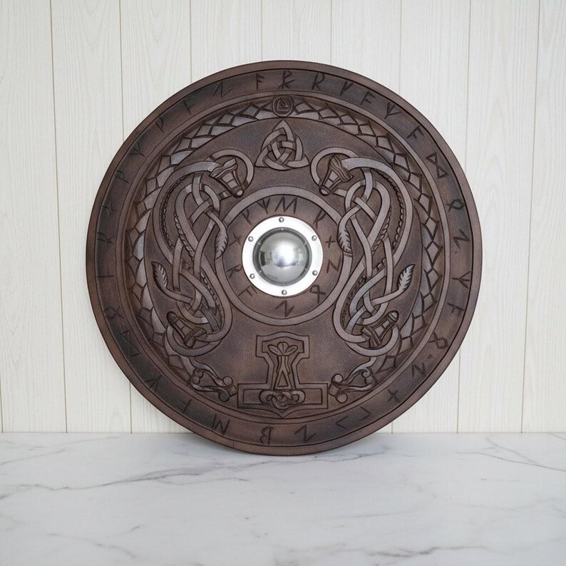 May include: A round, brown wooden shield with intricate carvings. The shield features a central silver boss, knotwork designs, and runes around the edge. A hammer symbol is carved at the bottom.
