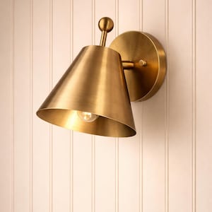 May include: A brass wall sconce with a conical shade and a round backplate. The sconce has a small bulb inside the shade and is mounted on a white vertical-paneled wall. The fixture is made of brass.