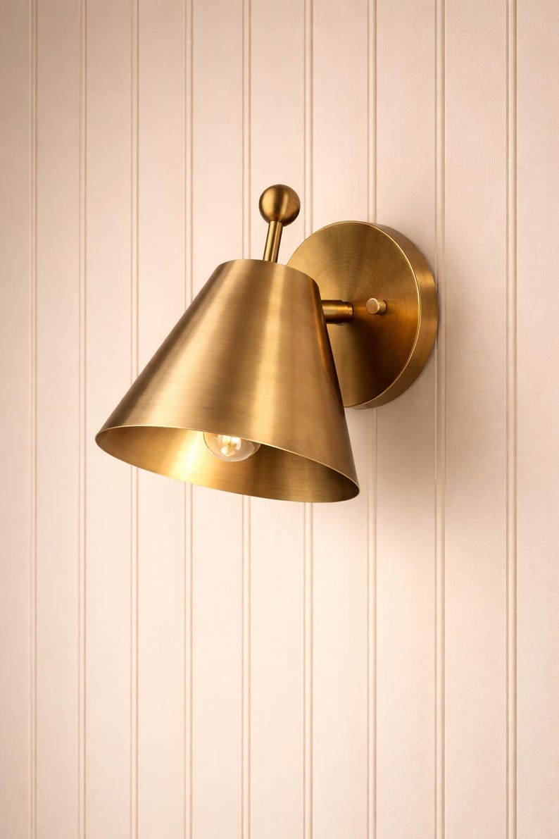 May include: A brass wall sconce with a conical shade and a round backplate. The sconce has a small bulb inside the shade and is mounted on a white vertical-paneled wall. The fixture is made of brass.