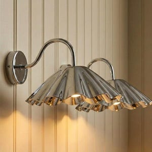 May include: Two wall-mounted light fixtures featuring polished silver-toned metal shades with a distinct scalloped, fluted edge design. Each lamp is attached to the wall by a circular base and a curved arm, emitting a warm glow from underneath.
