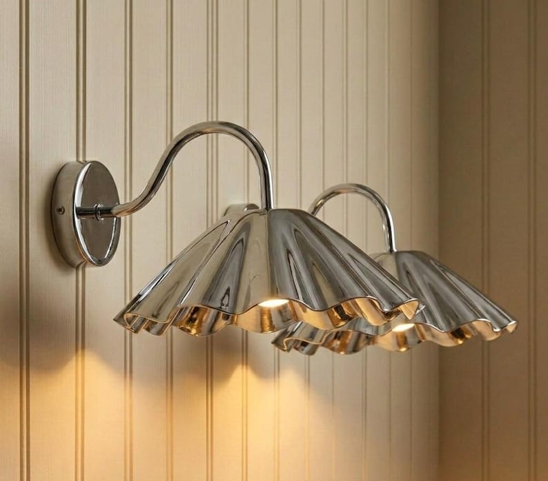 May include: Two wall-mounted light fixtures featuring polished silver-toned metal shades with a distinct scalloped, fluted edge design. Each lamp is attached to the wall by a circular base and a curved arm, emitting a warm glow from underneath.