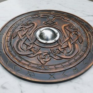 May include: A round, wooden shield with intricate carvings. The shield features a central metal boss, a hammer symbol, and intertwined serpent designs. The outer rim has etched runes. The shield is a dark brown color.