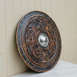 May include: A round, wooden shield with intricate carvings and a central metal boss. The shield features Norse-inspired designs, including a hammer symbol and runic inscriptions around the edge. The shield is primarily brown with black accents.