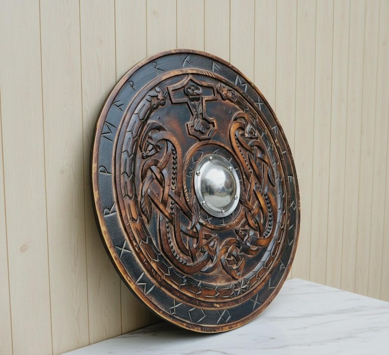 May include: A round, wooden shield with intricate carvings and a central metal boss. The shield features Norse-inspired designs, including a hammer symbol and runic inscriptions around the edge. The shield is primarily brown with black accents.