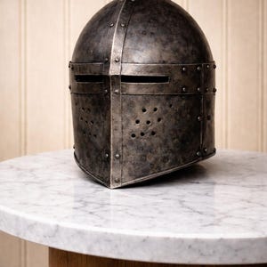 May include: A weathered, dark gray metal helmet with riveted construction and horizontal eye slits. The helmet features numerous small ventilation holes and sits on a white marble-topped table with a wooden base. The helmet's design suggests a medieval theme.