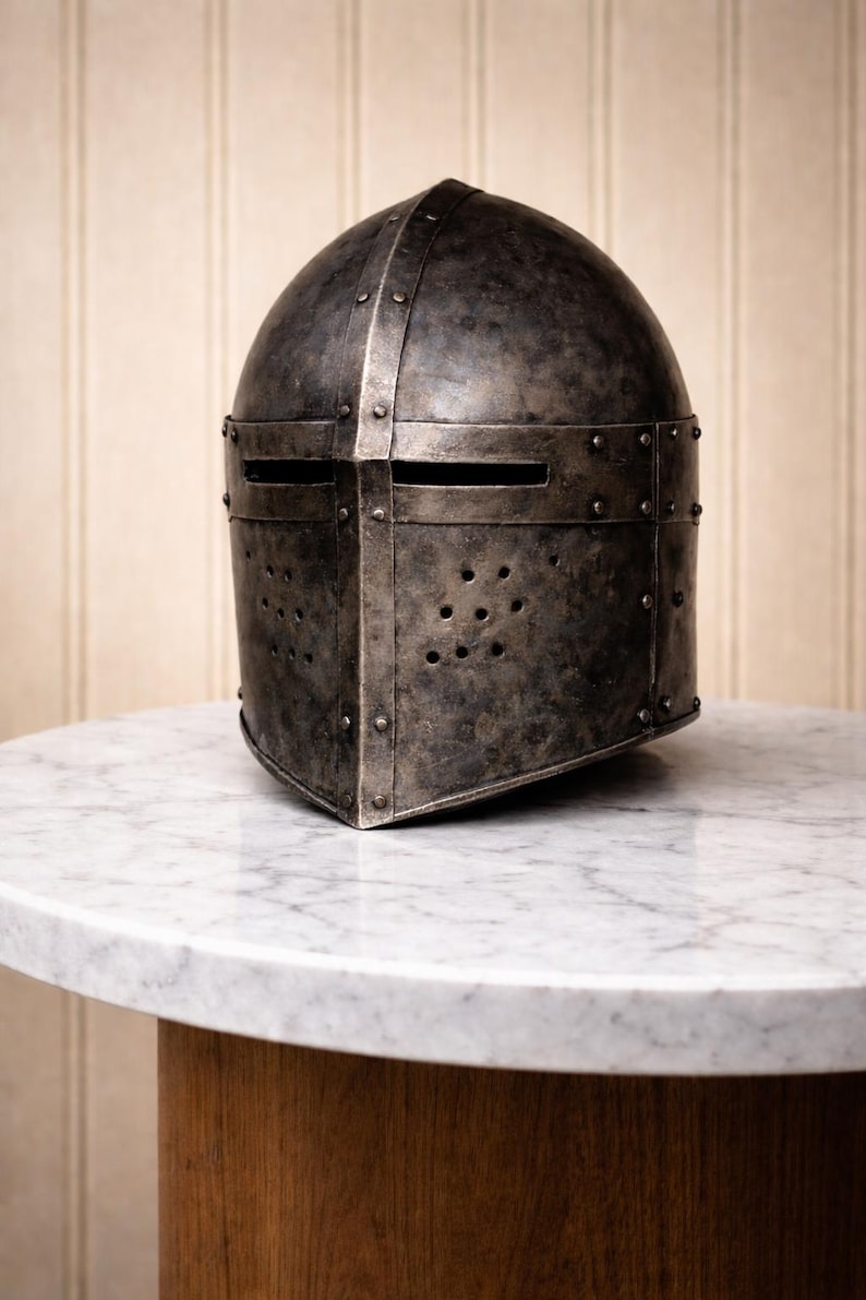 May include: A weathered, dark gray metal helmet with riveted construction and horizontal eye slits. The helmet features numerous small ventilation holes and sits on a white marble-topped table with a wooden base. The helmet's design suggests a medieval theme.