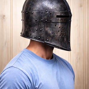 May include: A weathered, dark metal helmet with a visor and rivet detailing. The helmet has a rectangular opening for the eyes and several small holes below. The person is wearing a light blue t-shirt.