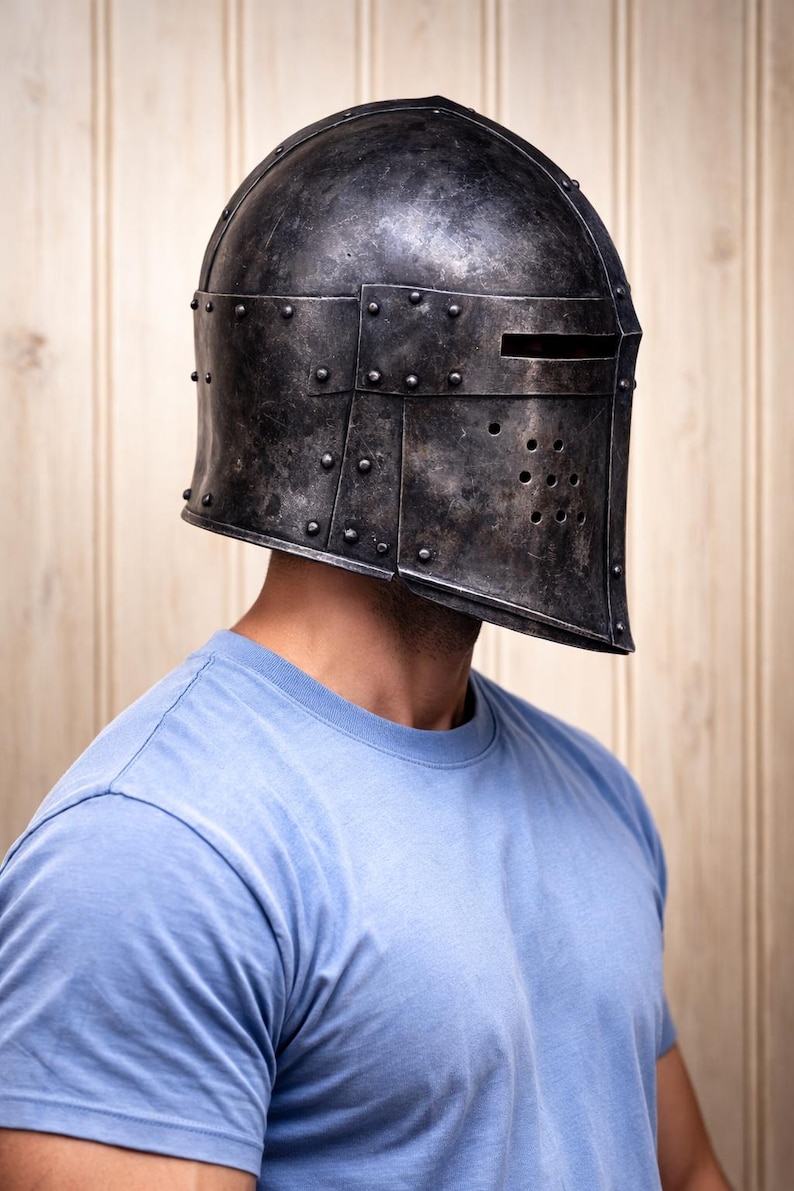 May include: A weathered, dark metal helmet with a visor and rivet detailing. The helmet has a rectangular opening for the eyes and several small holes below. The person is wearing a light blue t-shirt.