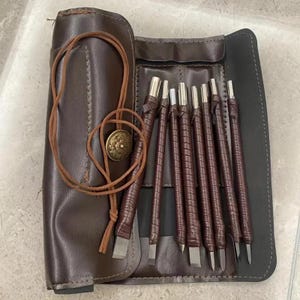 May include: A set of seven wood carving tools with brown leather-wrapped handles and metal tips, organized in a dark brown leather roll-up case. The case features a long brown leather cord and a circular brass-toned button for secure closure.