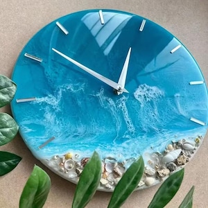 May include: Circular wall clock with a blue resin face showing an ocean design. The clock has white hands and silver markers. The lower edge is adorned with seashells and small stones. A distinctive decorative item.