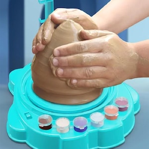 May include: A bright turquoise plastic pottery wheel toy featuring a rotating base and a built-in tray holding seven small paint pots in various colours. The device includes a vertical support arm and is shown with a mound of wet brown clay.