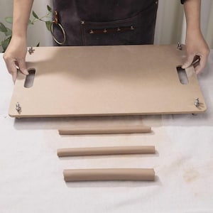 May include: A light brown wooden craft board with two handle cutouts and metal fasteners. Three rolled brown paper tubes are arranged below the board on a white surface. The board is held by a person wearing a brown apron.