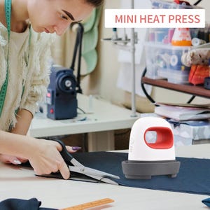 May include: A white and red mini heat press with a gray base sits on a table next to a pair of scissors and a dark blue fabric. The text "MINI HEAT PRESS" is displayed in the upper right corner.
