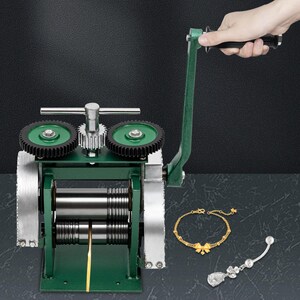 May include: A green metal rolling mill with silver rollers and gears. A hand turns the handle. A gold bracelet and a silver earring with a floral design are displayed next to the mill.