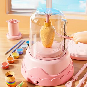 May include: A pink pottery wheel kit featuring a clear protective dome, a central rotating platform holding a yellow clay vase, a set of small paint pots in various colours, two paint brushes, and sculpting tools on a light wooden surface.