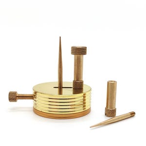 May include: A set of brass tools, including a round base with a vertical spike, a threaded adjustment screw, and several removable components. The tools have a polished gold finish and are designed for a specific purpose.