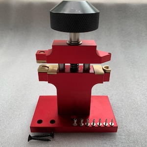 May include: A red metal watch case press tool featuring a black knurled adjustment knob at the top, a central spring-loaded mechanism with brass inserts, and a flat base with several small metal pins and two separate black screws included.