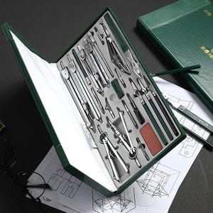 May include: A green leatherette case with a variety of silver drafting tools, including compasses, dividers, and rulers. The tools are neatly arranged in a gray foam insert. A green notebook and architectural drawings are also visible.