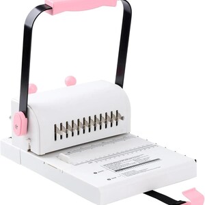 May include: A white paper binding machine featuring a black metal handle with a pink grip. The device includes a white base with printed measurement scales in centimetres and inches, a row of metal punching teeth, and pink adjustment knobs.