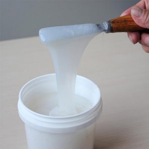 May include: A white bucket filled with a viscous, translucent substance. A metal spatula with a wooden handle holds a portion of the substance, which is dripping down. The image suggests a crafting or industrial material.