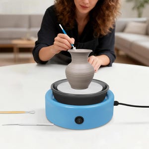 May include: A light blue electric pottery wheel with a black basin and a central white disc supporting a grey clay vase. A blue-handled tool is held above the vase, while two metal sculpting tools lie on the surface nearby for ceramic work.