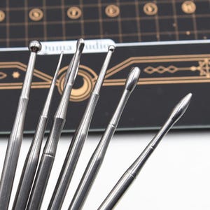 May include: A collection of silver-colored metal tools with various tip shapes, including rounded, pointed, and angled designs. The tools are arranged against a dark background with gold accents and text.