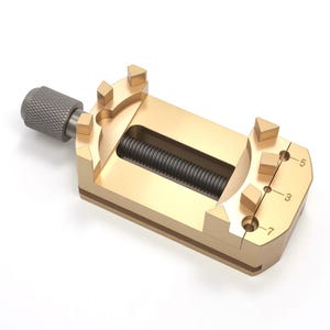 May include: A gold-colored vise with a threaded rod and a textured knob. The vise has a rectangular body with a central channel and is marked with the numbers 3, 5, and 7. This tool is used for holding objects securely.