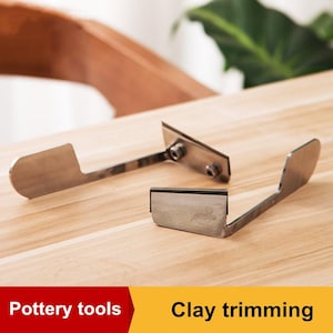 May include: Two stainless steel pottery tools for clay trimming are displayed on a light wood surface. The tools have a blade and a handle, with one tool featuring a serrated edge. The image also includes the text "Pottery tools" and "Clay trimming".