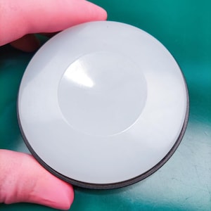 May include: A white, circular object with a black rim. The object has a slightly raised, circular center. The item is held by a person's hand, and the background is a solid green color.