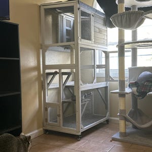May include: A multi-level cat enclosure with a wooden frame and wire mesh panels. The enclosure has multiple levels, doors, and a small house-like structure on top. It sits on wheels for easy movement. A cat tree is visible in the background.