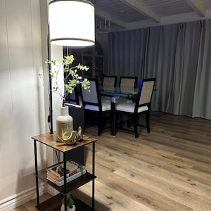 May include: A modern living room scene featuring a floor lamp with a white shade, a three-tiered side table, and a dining table. The side table has a dark wood finish and holds books, a vase, and decorative items. The dining table has a glass top and is surrounded by black chairs.