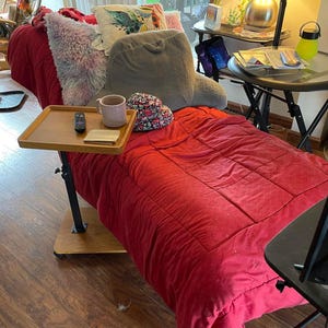 May include: A red quilted blanket is the focal point of this image, complemented by various pillows in shades of pink, gray, and patterned designs. A wooden tray table holds a remote, a mug, and a small notepad. The scene suggests a cozy, comfortable living space.