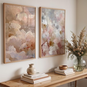 Puede incluir: Dos impresiones artísticas abstractas enmarcadas con pinceladas en forma de nube en tonos rosa pálido, melocotón y blanco. La obra de arte se exhibe en una pared blanca sobre una consola de madera.