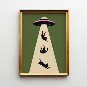 Op de afbeelding: Een ingelijste kunstprint met een humoristische scène: een zwart-roze UFO die drie zwarte katten omhoog straalt. De achtergrond is olijfgroen. De katten zijn als silhouetten in een lichtstraal.