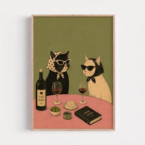 May include: A framed art print featuring a black cat wearing a polka dot headscarf and white sunglasses, and a cream cat wearing a black headscarf and black sunglasses. They sit at a pink table with wine, cheese, olives, and a book titled Nine Lives.