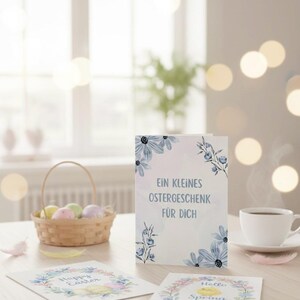 K&ouml;nnte beinhalten: Ein Stillleben mit Ostergru&szlig;karten und Dekorationen. Eine Karte mit dem Text "Ein kleines Ostergeschenk f&uuml;r Dich" wird zusammen mit einem Korb mit pastellfarbenen Eiern und einer Tasse Kaffee gezeigt.