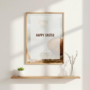 K&ouml;nnte beinhalten: Ein gerahmter Druck mit dem Text "HAPPY EASTER" in braunen Buchstaben. Der Druck zeigt eine neutrale Farbpalette mit Osterei-Bildern. Eine kleine Topfpflanze und eine Vase mit Zweigen befinden sich auf einem Holzregal darunter.