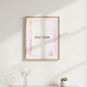 K&ouml;nnte beinhalten: Ein gerahmter Druck mit den Worten "HELLO SPRING" in einer weichen, rosa Schriftart. Das Kunstwerk zeigt ein Design im Aquarellstil mit rosa und orangen Farbt&ouml;nen. Eine kleine Blume befindet sich in der unteren rechten Ecke. Eine Vase mit wei&szlig;en Blumen und eine kleine Sukkulente stehen auf einem Regal.