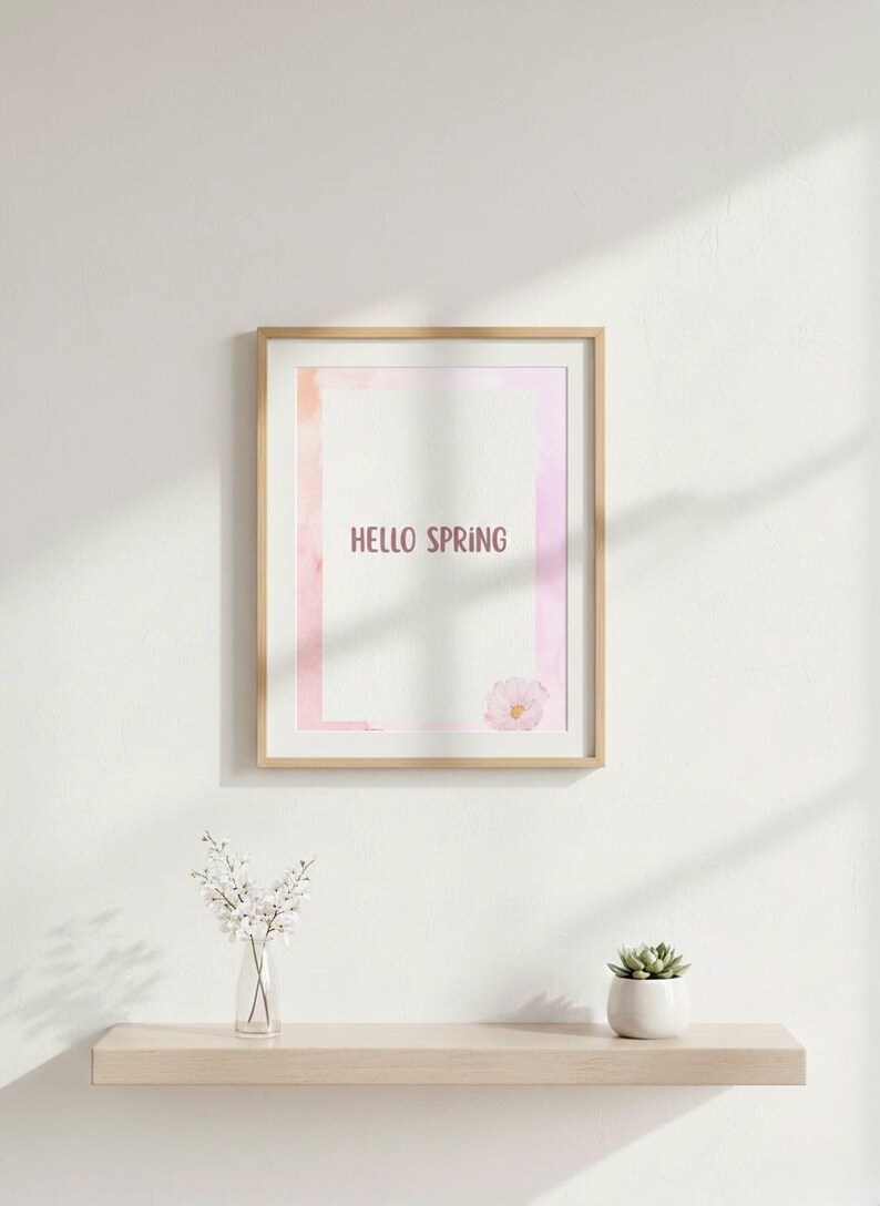K&ouml;nnte beinhalten: Ein gerahmter Druck mit den Worten "HELLO SPRING" in einer weichen, rosa Schriftart. Das Kunstwerk zeigt ein Design im Aquarellstil mit rosa und orangen Farbt&ouml;nen. Eine kleine Blume befindet sich in der unteren rechten Ecke. Eine Vase mit wei&szlig;en Blumen und eine kleine Sukkulente stehen auf einem Regal.