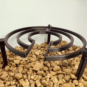 Könnte beinhalten: Ein dunkelbrauner, 3D-gedruckter Pflanzenständer mit kreisförmigem Design. Der Ständer hat drei Beine und eine Reihe konzentrischer Ringe. Der Ständer ist für einen Blumentopf konzipiert und steht auf einem Bett aus kleinen, hellbraunen Steinen.