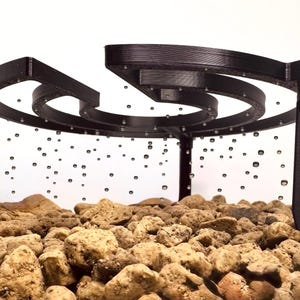 Könnte beinhalten: Ein schwarzes, spiralförmiges Wasserspiel mit einer Reihe konzentrischer Ringe, die von vier vertikalen Pfosten getragen werden. Das Design befindet sich über einem Bett aus kleinen, hellbraunen Steinen. Wassertröpfchen scheinen in der Luft zu schweben.