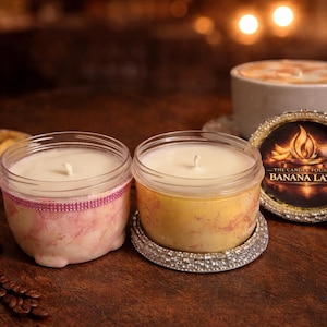 Può includere: Due candele in barattolo con cera bianca, una con un design marmorizzato rosa e l'altra con un design marmorizzato giallo. Le candele poggiano su basi decorative. Sullo sfondo una tazza di caffè e un piatto di fette di banana. L'etichetta recita "Banana Latte".