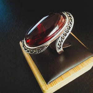 Puede incluir: Un anillo de plata con una gran piedra preciosa ovalada de color rojo intenso. El anillo tiene una banda decorativa con pequeños acentos redondos de color plateado. El anillo se exhibe sobre una superficie marrón oscuro con una base de madera marrón claro.