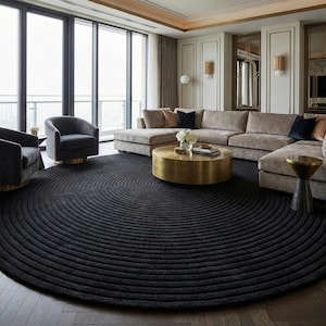 May include: A large, round black rug with a concentric ripple design dominates the living room. The rug sits on a hardwood floor, complementing the neutral-toned sectional sofa and gold accents. Two gray armchairs and a gold coffee table complete the modern decor.